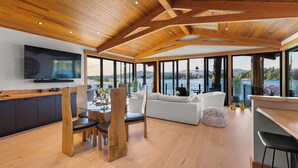A spacious living area with a wooden ceiling, a large TV mounted on the wall, a white sofa, and a dining table with chairs. - Kingfisher Manor (Sooke)