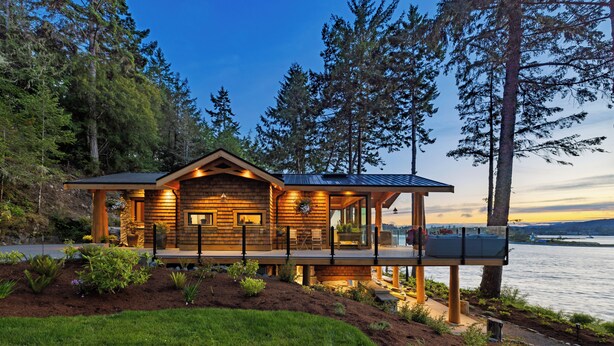 A wooden cabin with a deck, surrounded by trees and a lake. - Kingfisher Manor (Sooke)