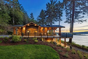 A wooden cabin with a deck, surrounded by trees and a lake. - Kingfisher Manor (Sooke)