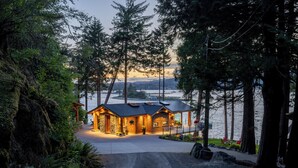 A wooden cabin with a stone foundation, surrounded by tall trees and a view of a lake at dusk. - Kingfisher Manor (Sooke)