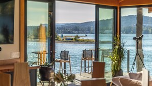 A room with large windows overlooking a lake, featuring a wooden ceiling, a potted plant, and two chairs. - Kingfisher Manor (Sooke)