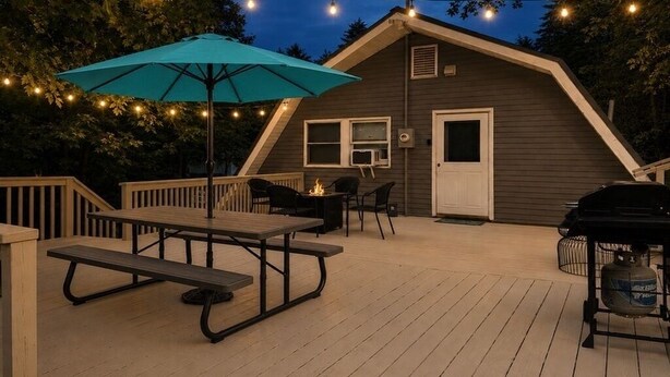Una terraza de madera con mesas y sillas de picnic, una parrilla y luces colgantes.