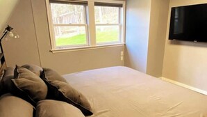 Un dormitorio con cama, almohadas, ventana y televisor.