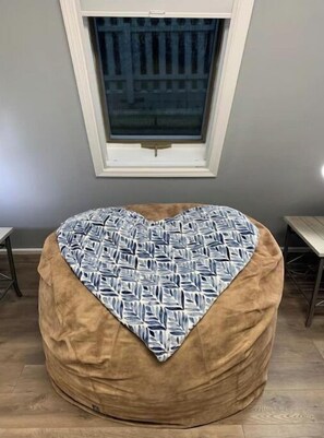 Sydämenmuotoinen papupussi, jossa on lehtikuviollinen koristetyyny, ja joka on sijoitettu huoneeseen, jossa on ikkuna ja puulattia.
