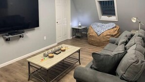 Una sala de estar con un televisor de pantalla plana, un sillón informal, un sofá gris y una mesita con una lámpara.