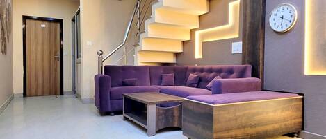 Un moderno vestíbulo de hotel con un sofá seccional púrpura, una mesa de centro de madera y una escalera con peldaños iluminados.