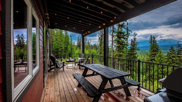 A wooden deck with a picnic table and chairs, overlooking a forest and mountains.