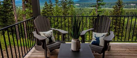 Een houten terras met twee Adirondack stoelen, een bijzettafeltje en een vaas met groen.