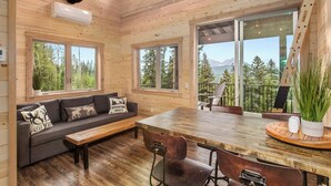 A cozy cabin interior with a wooden ceiling, a sofa with decorative pillows, a dining table with chairs, and large windows offering a view of trees and mountains.
