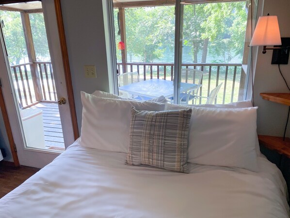 A bedroom with a bed, pillows, a window with a view of a deck and trees, and a table with chairs outside.