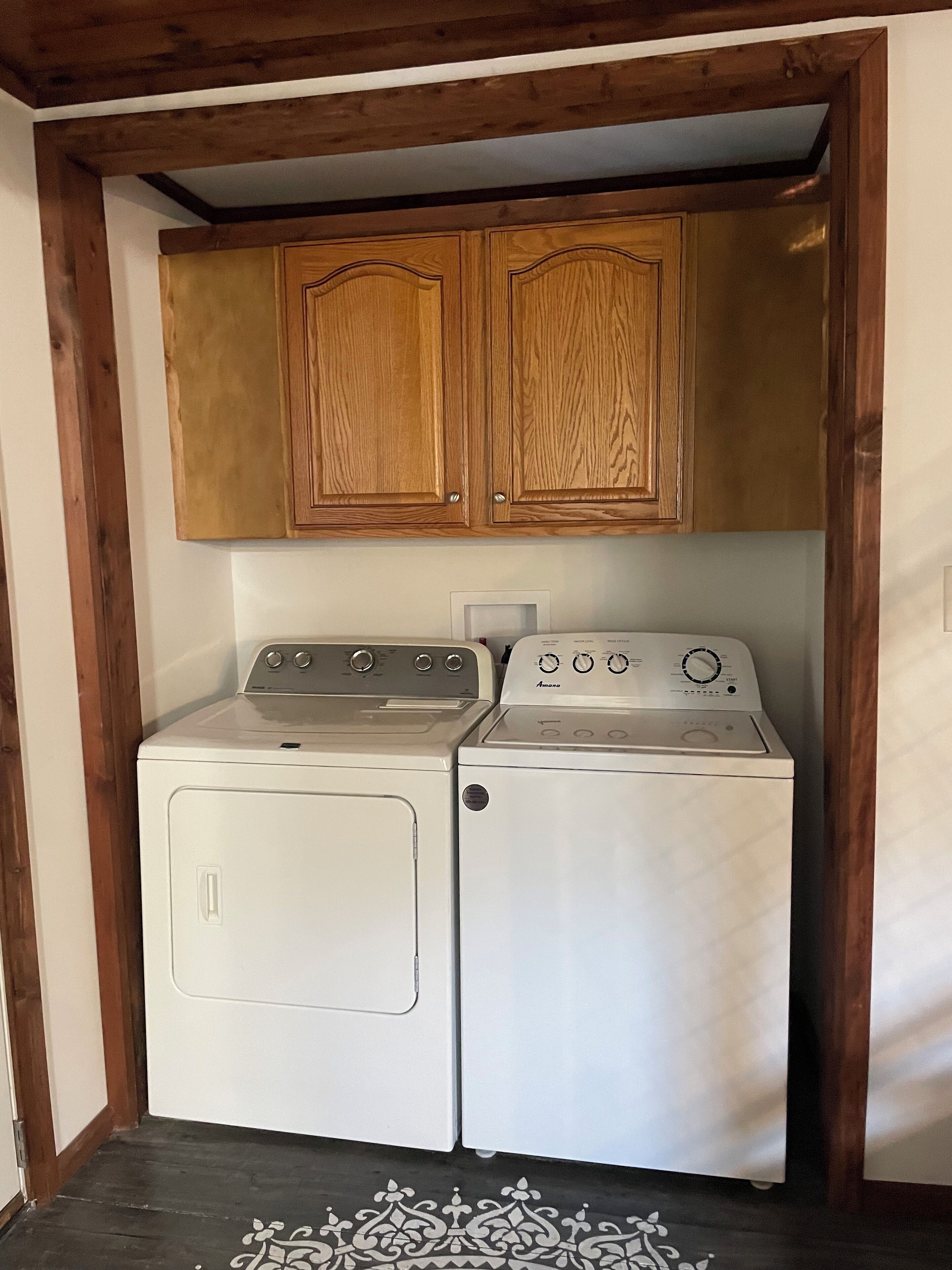 A laundry room with two white front-loading washers and dryers, wooden cabinets above, and a patterned rug on the floor.