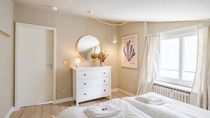 Ett sovrum med två sängar, en vit byrå, en rund spegel, en vägglampa och ett fönster med gardiner.
