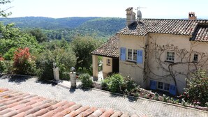 Ein Gebäude mit einem Ziegeldach, ein Garten mit verschiedenen Pflanzen und Blick auf eine hügelige Landschaft.
