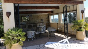 Eine Terrasse im Freien mit Steinboden, Holzbalken, Korbmöbeln und Topfpflanzen.