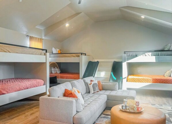 Un moderno loft con un sofá gris, una mesa redonda de madera y dos literas con ropa de cama naranja.