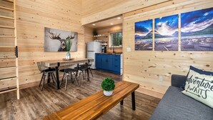 Ein gemütliches Blockhaus-Interieur mit einer Essnische aus Holz, einer Küche mit blauen Schränken und einem Wohnbereich mit grauem Sofa und einem Wandbild von einer Berglandschaft.