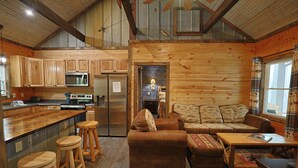 Interior rústico de una cabaña con paredes de madera, una cocina con electrodomésticos modernos, una zona de estar acogedora con sofá y reposapiés, y una barra con taburetes.