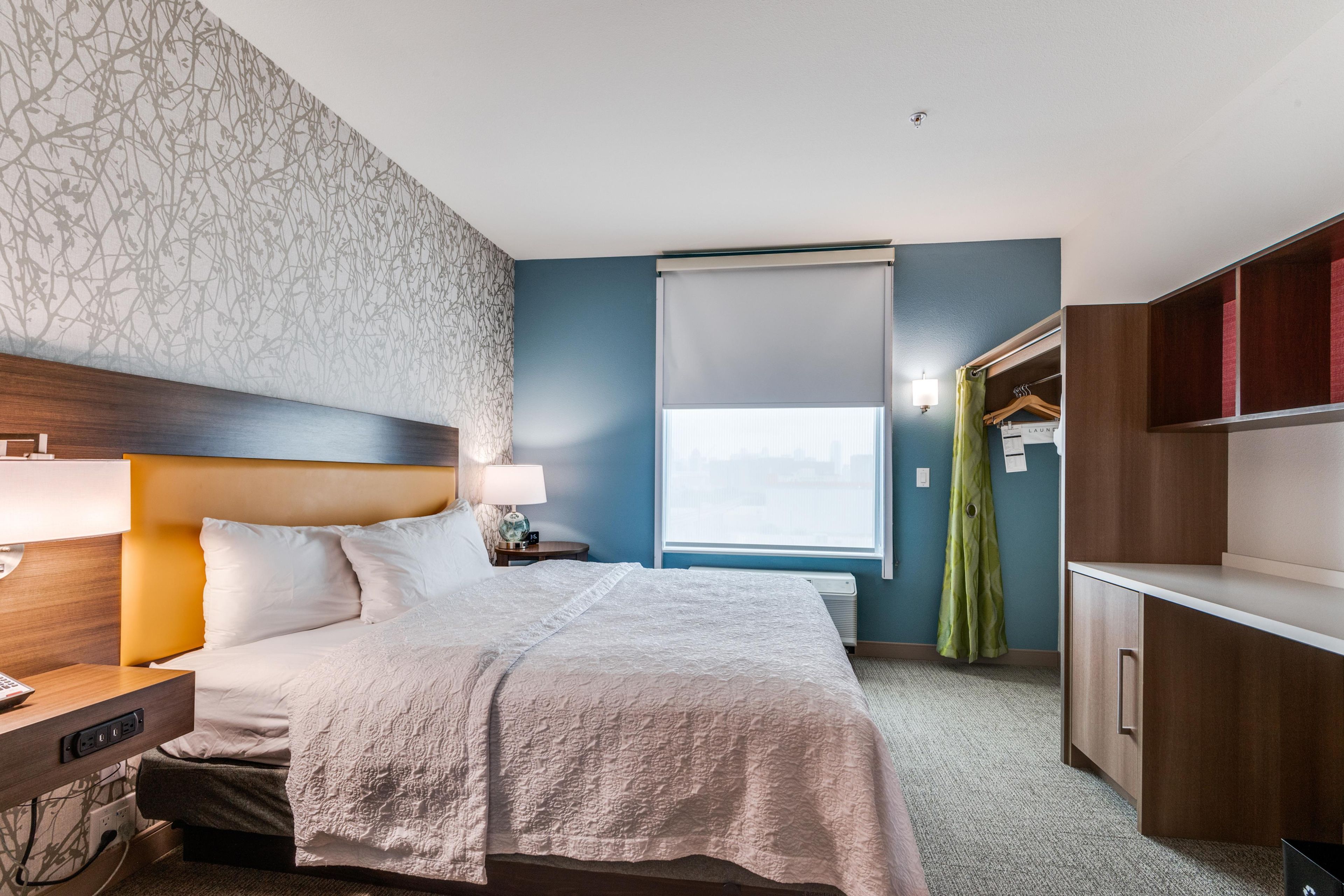 A hotel room with a bed, bedside table, lamp, window with blinds, and a wardrobe.