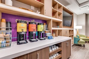 Un espace dédié au café, équipé de plusieurs distributeurs de café étiquetés « GOURMET », « EAU CHAUDE » et « CAFÉ DÉCAFÉINÉ ». On y trouve également des étagères remplies de tasses et un mur orné d’une fresque sur le thème du café.