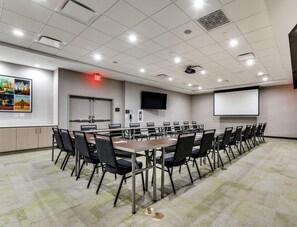 Une salle de conférence avec une longue table, des chaises, un écran de projection, un téléviseur et un panneau indiquant la sortie.