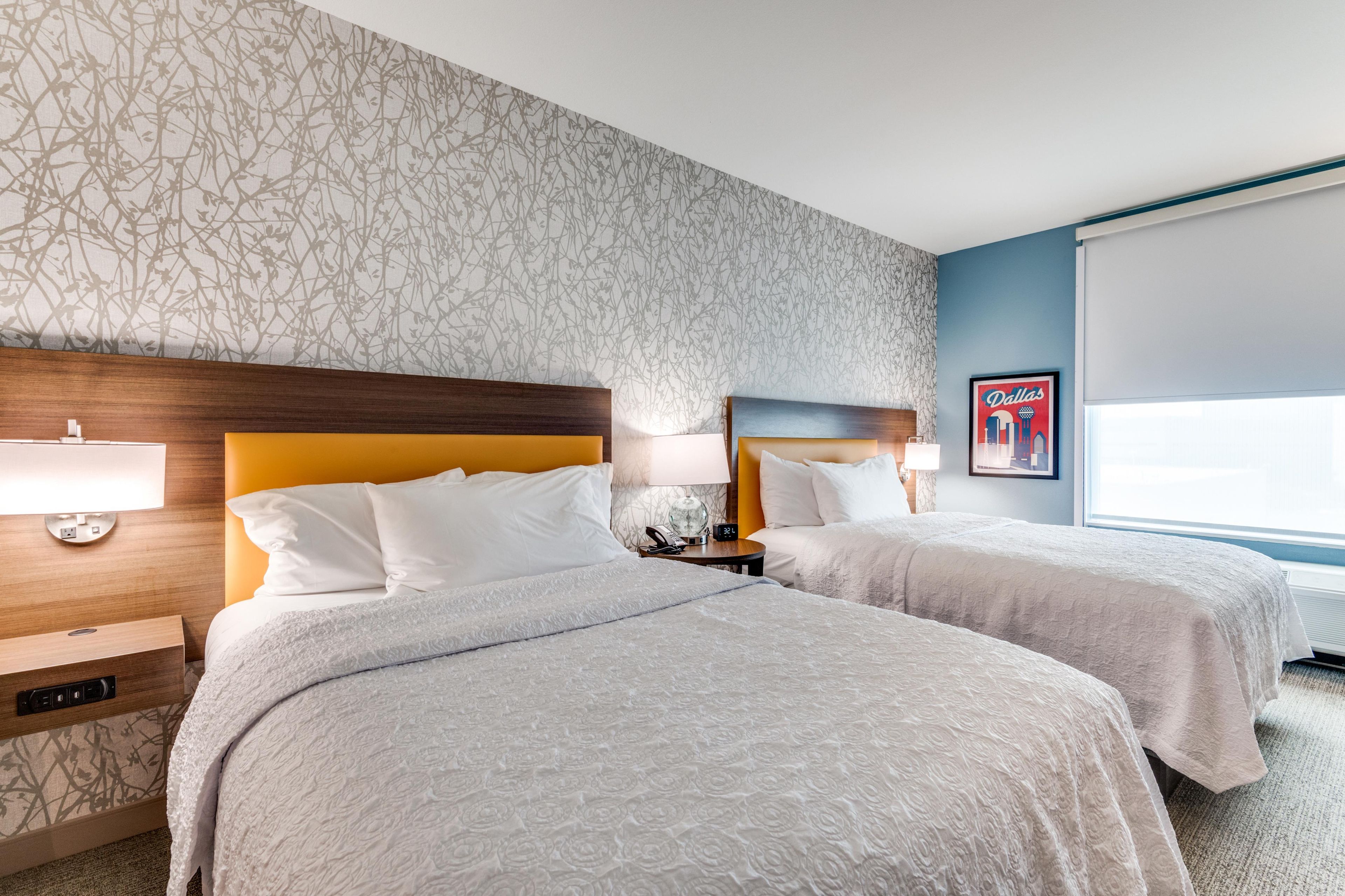 A hotel room with two beds, a nightstand, a lamp, a framed picture on the wall, and a window.