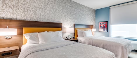 Et hotelværelse med to senge, et natbord, en lampe, et billede i ramme på væggen og et vindue.