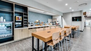 Ein modernes Café mit einem langen Holztisch und Stühlen, einer Kühlvitrine und einer Auswahl an Speisen und Getränken.