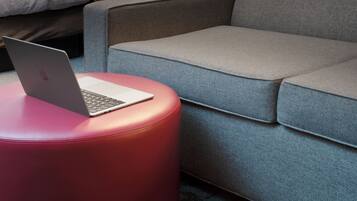 A modern hotel room with a grey sofa, a red ottoman with a laptop, a bed with white bedding, and a geometric-patterned wall.