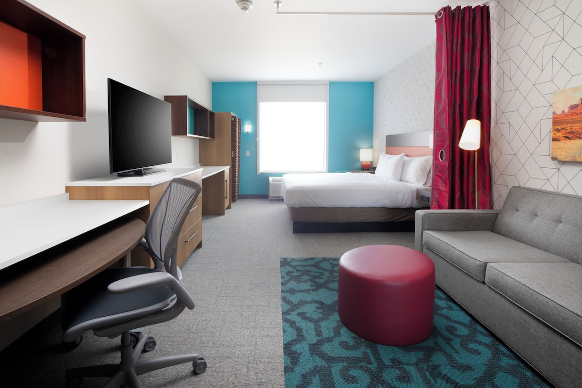 Ein Hotelzimmer mit Bett, Sofa, Schreibtisch mit Stuhl, Fernseher und einem fenster mit Vorhängen.