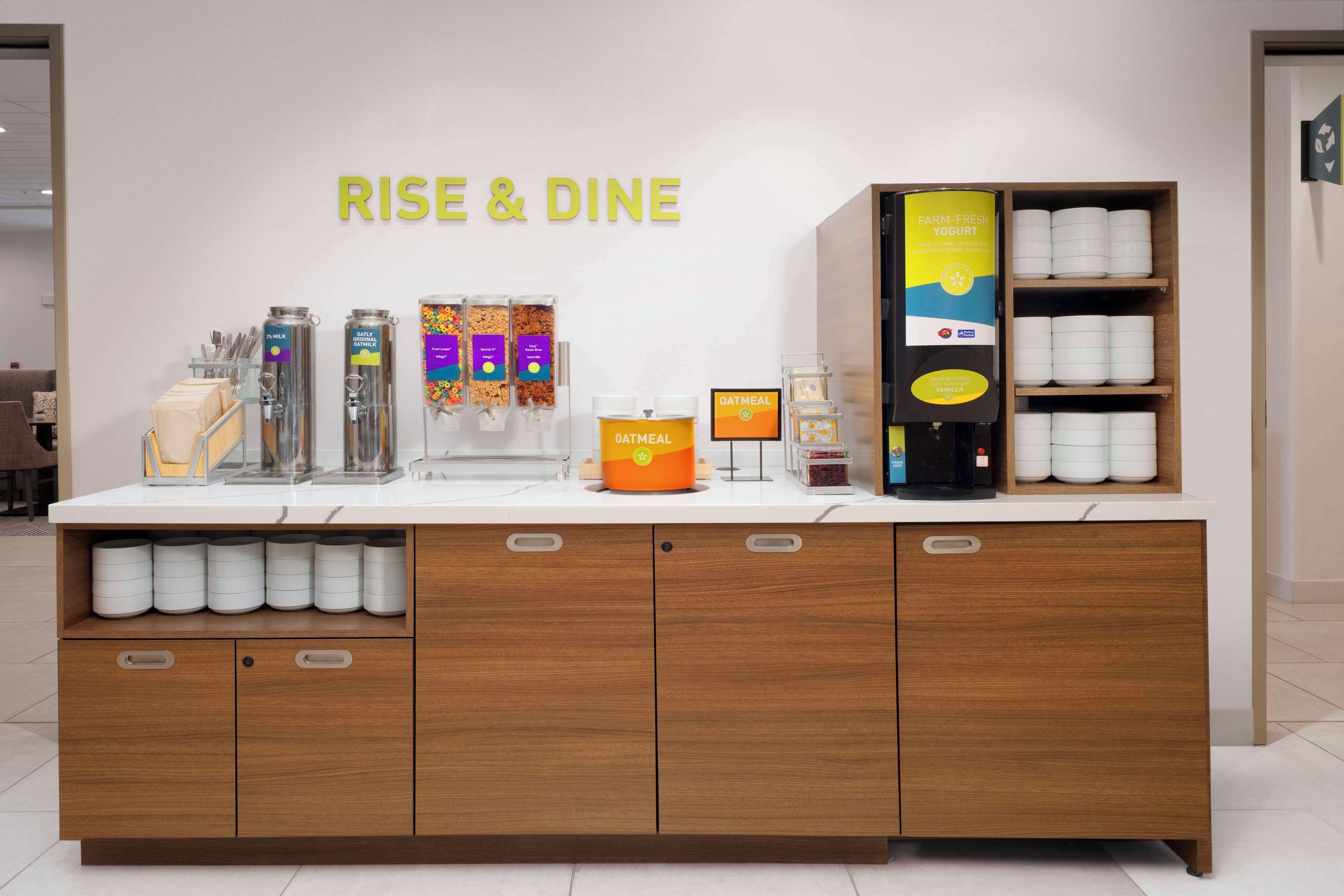 A breakfast bar with a sign reading "RISE & DINE," featuring a coffee machine, various food containers, and a shelf with bowls and cups.