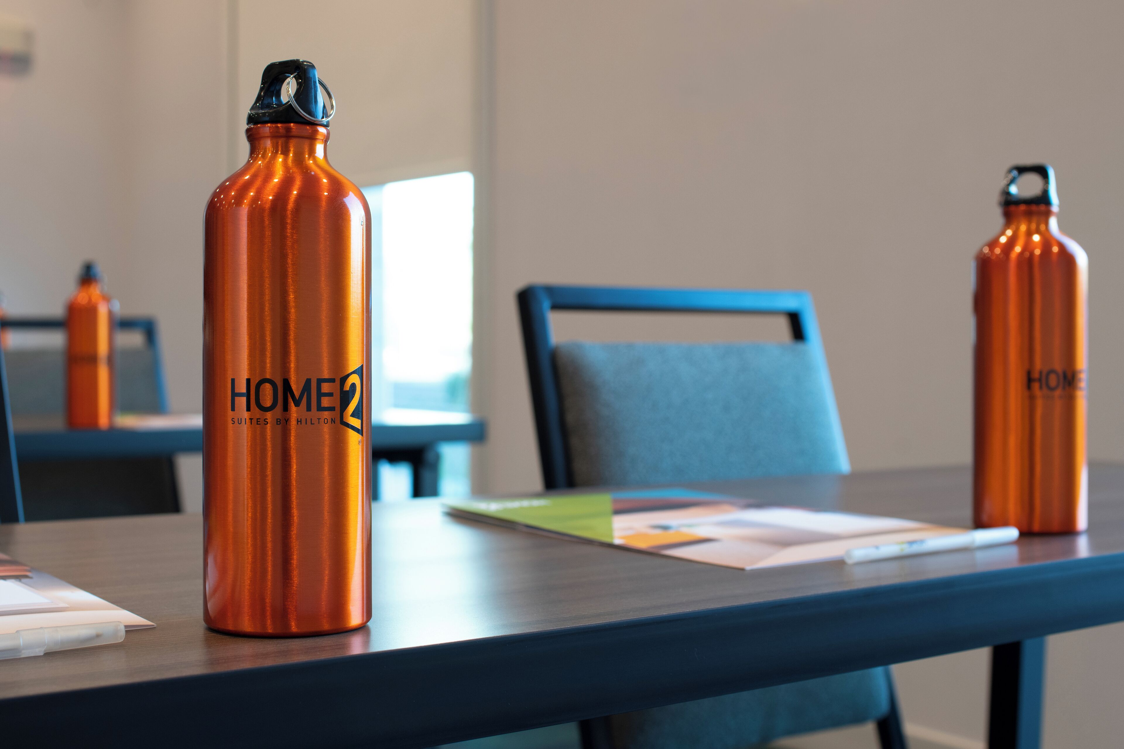 A hotel room with a wooden table, two orange water bottles labeled "HOME2 SUITES BY HILTON", a blue chair, and a window in the background.