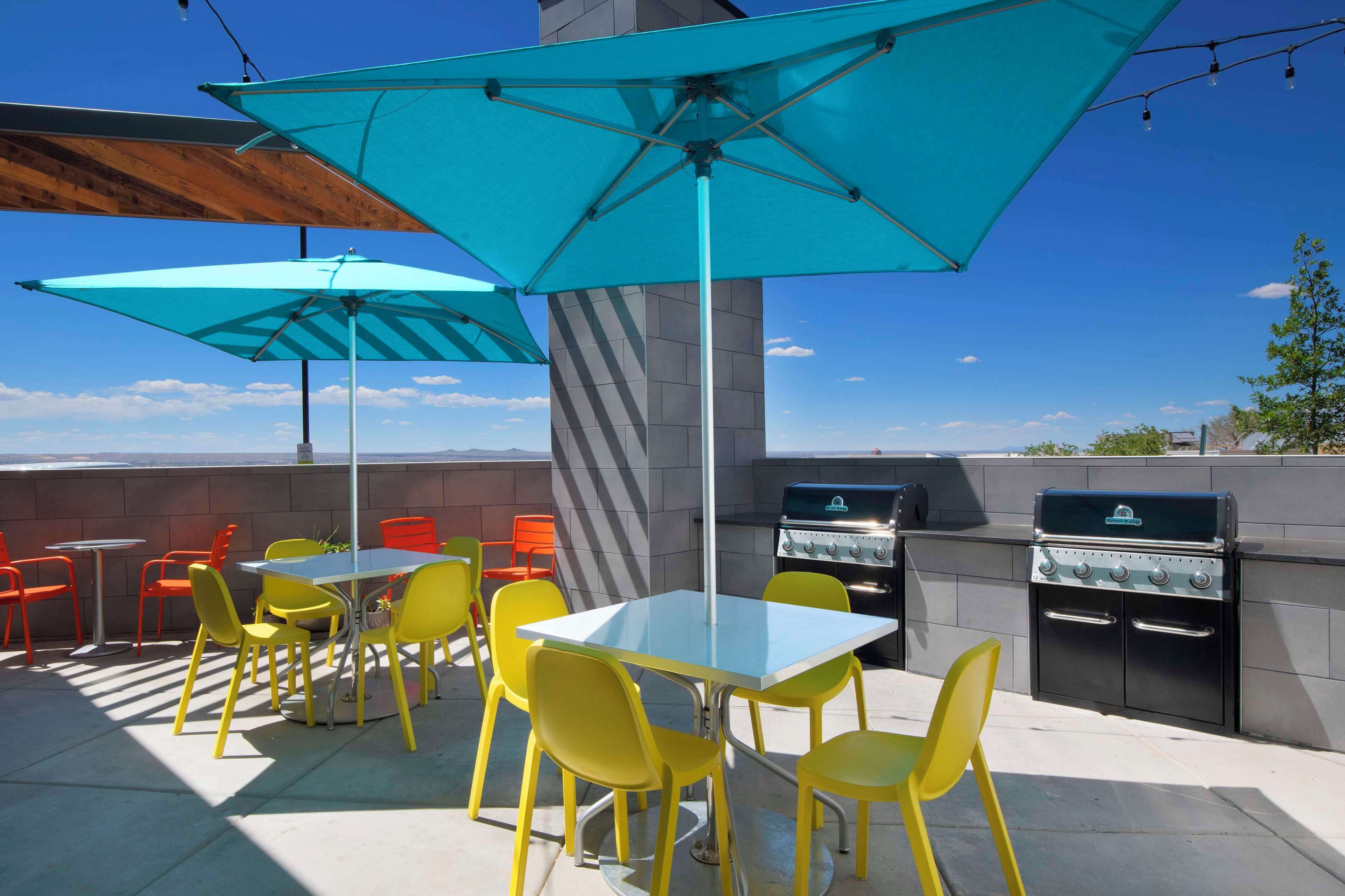 An outdoor patio with yellow chairs and blue umbrellas, a grill, and a view of the sky.
