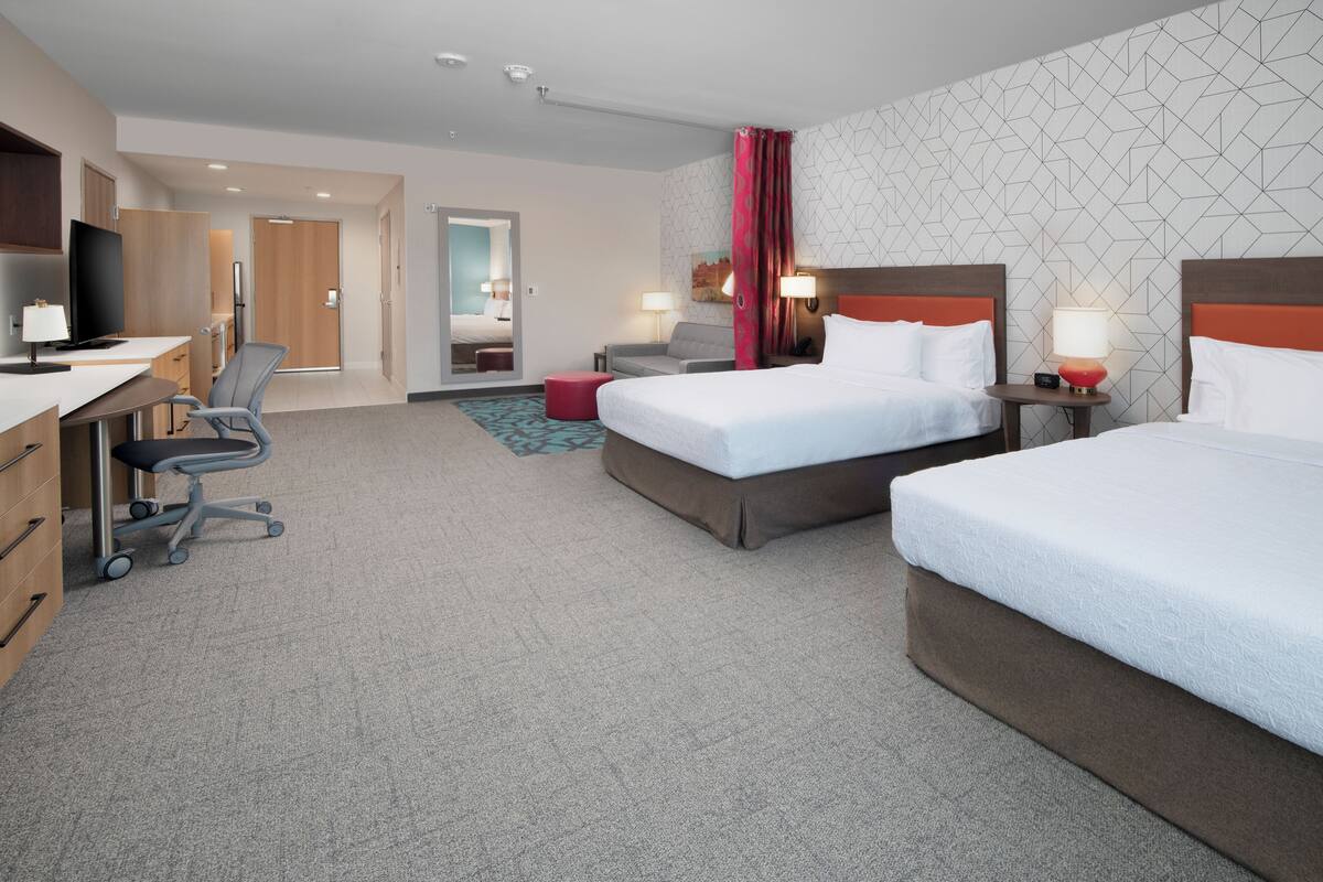 Ein Hotelzimmer mit zwei Betten, einem Schreibtisch mit Stuhl, einem Fernseher und einem kleinen Tisch mit Lampe.