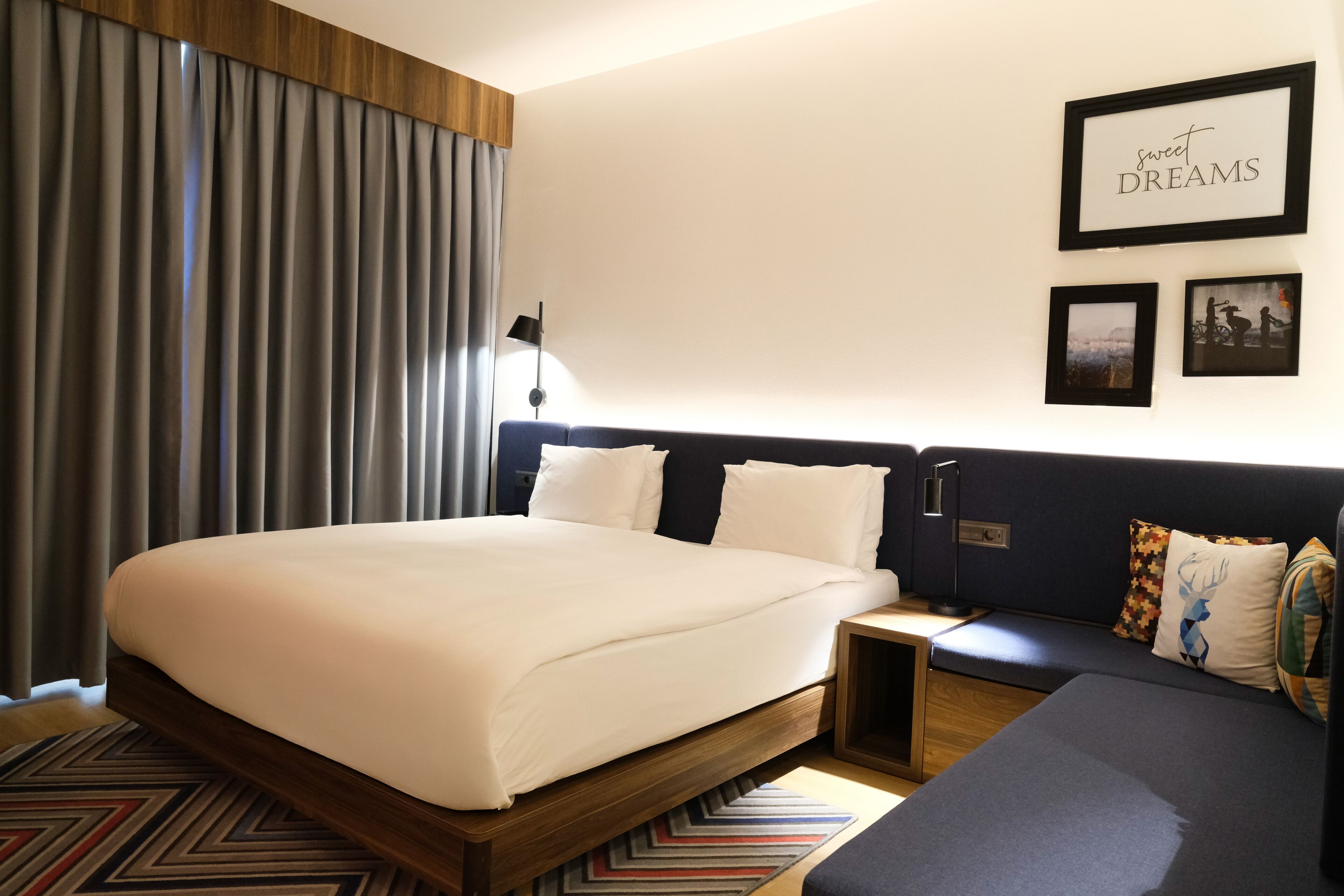 A modern hotel room with a large bed, a sofa, a nightstand, and a wall with framed pictures.