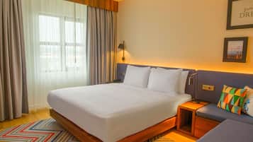 Habitación de hotel con una cama grande, una mesita de noche, un pequeño sofá y vistas al exterior a través de cortinas traslúcidas.
