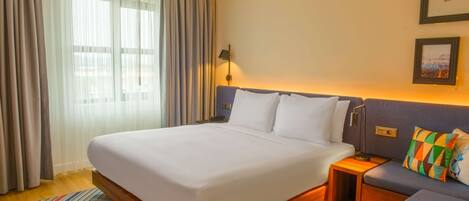 Habitación de hotel con una cama grande, una mesita de noche, un pequeño sofá y vistas al exterior a través de cortinas traslúcidas.