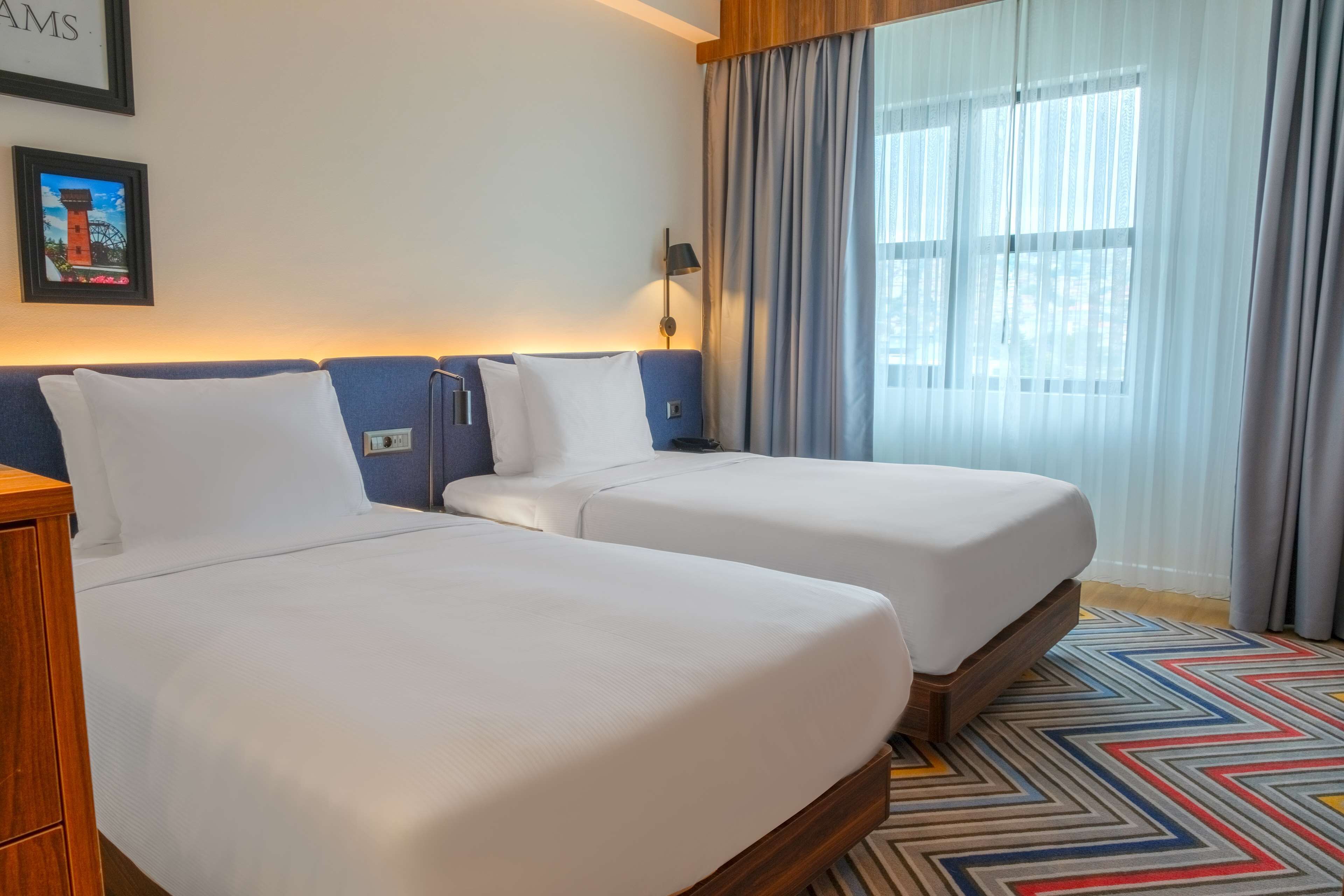 A hotel room with two beds, a wooden nightstand, a lamp, a framed picture on the wall, and a window with curtains.