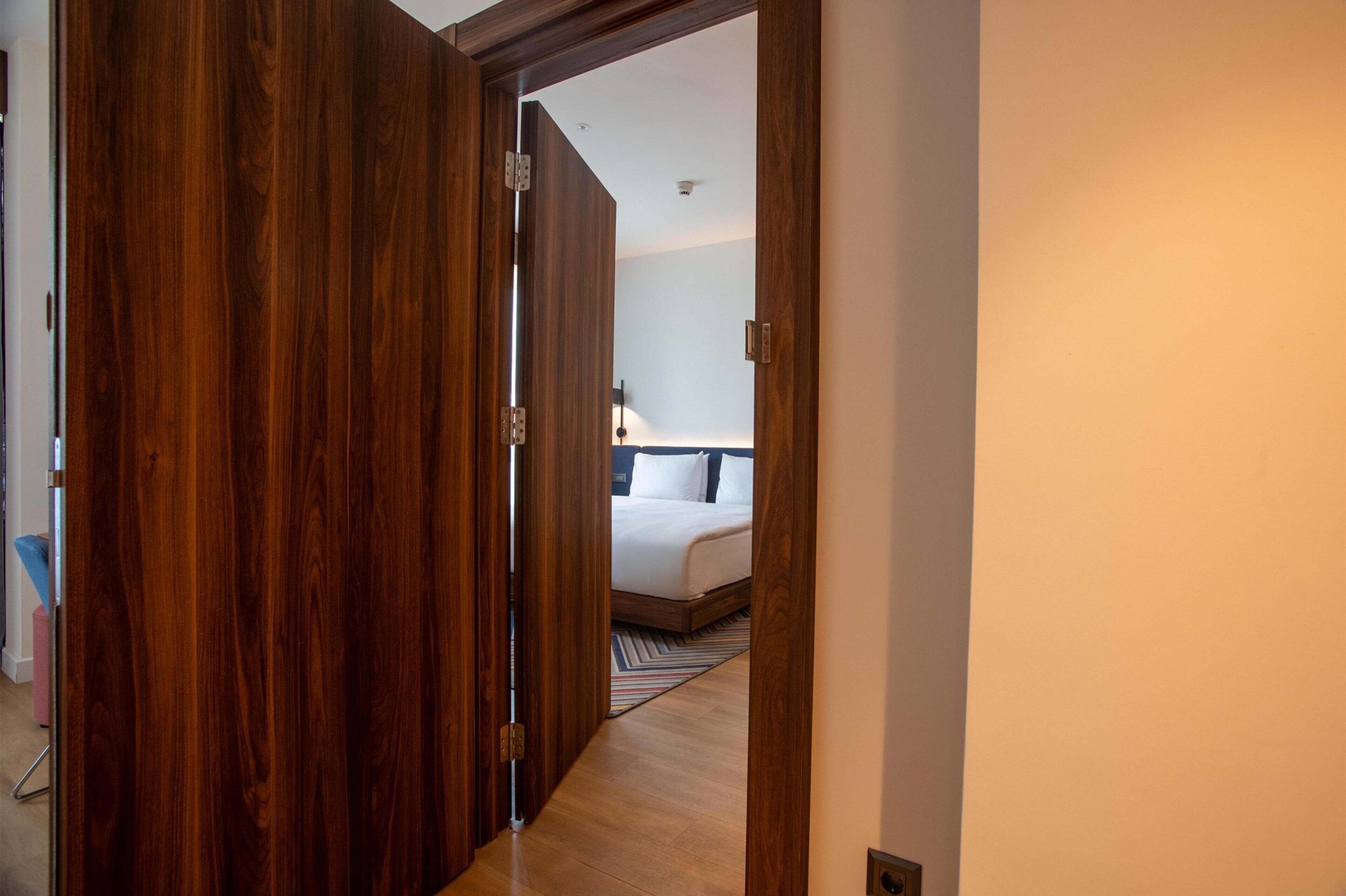 A hotel room with a wooden door, a bed with white bedding, and a striped rug on the floor.