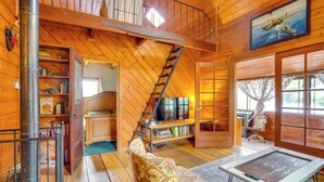 L’intérieur d’un chalet en bois comprend une chambre à coucher située en mezzanine, une cheminée, un téléviseur et une grande fenêtre.