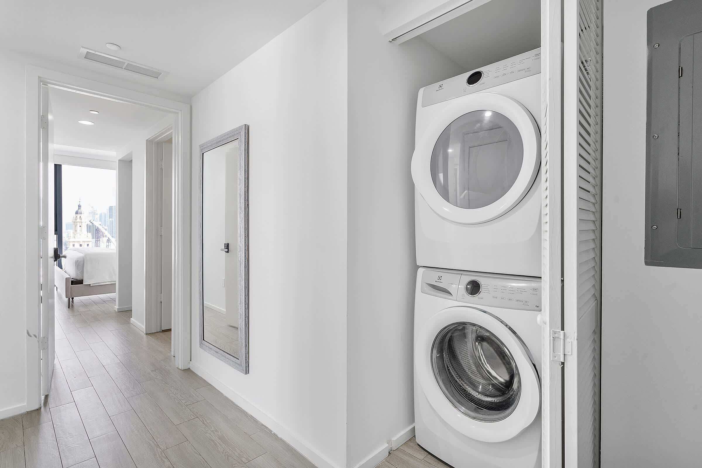 Une buanderie équipée d’un lave-linge et d’un sèche-linge, d’un grand miroir et offrant une vue sur le paysage urbain grâce à une porte ouverte.