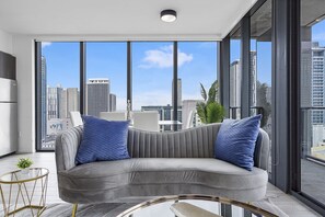 A modern living room with a grey sofa, two blue pillows, a glass coffee table, and a view of a cityscape through large windows.