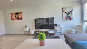 A modern living room with a flat-screen TV, a white coffee table with a green plant, and a sofa. There are two white chairs and a small white stool.