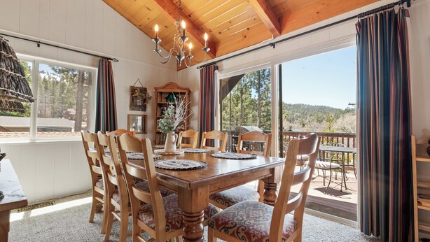 Un comedor con mesa y sillas de madera, una araña y ventanas grandes con cortinas.