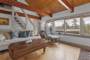 Un salón amplio con techo de madera, una escalera, un ventanal con vistas a árboles, un sillón estampado y un sofá blanco con cojines.
