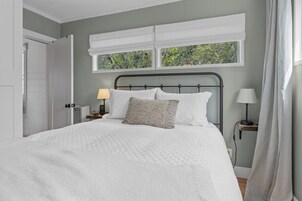 Una cama bien tendida con colcha blanca, una lámpara de noche y una ventana con vistas a la vegetación.