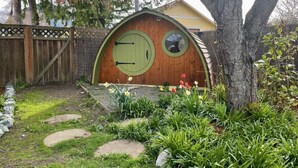 Una acogedora casa de madera estilo hobbit con puerta circular y pequeña ventana, rodeada de un jardín con piedras para pisar y diversas plantas.