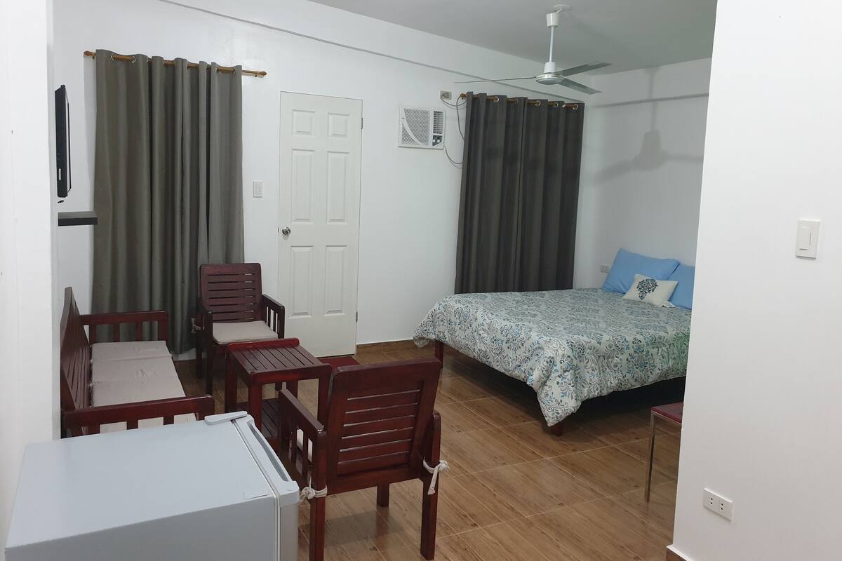 A bedroom with a bed, a desk, two chairs, a ceiling fan, and an air conditioning unit.