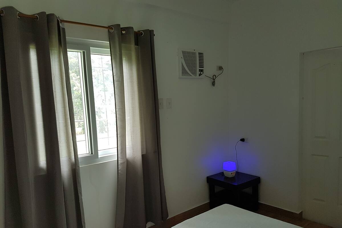 A bedroom with a bed, a window with curtains, a wall-mounted heater, and a bedside table with a lamp.