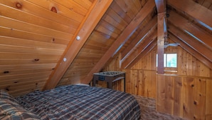 A cozy attic bedroom with wooden walls, a bed with plaid bedding, and a foosball table. - Far Enough Cabin (Sugarloaf)