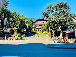 A street view with a building that has multiple floors and a sloped roof. There are trees and a clear blue sky. - EXCELENTE OPÇAO - 03 quadras RUA COBERTA (Gramado)
